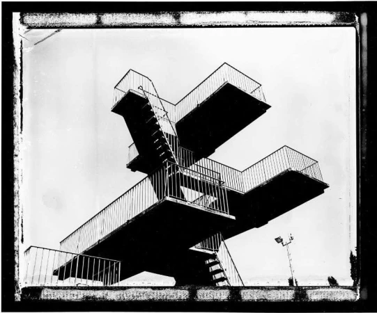 Monochrome photograph of a high diving platform with stairs and railings against a bright sky, showcasing its architectural structure.