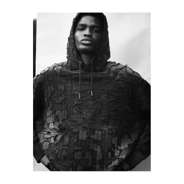 Monochrome studio portrait of a Black male model in a distressed hoodie, gazing directly into the camera against a bright white background.