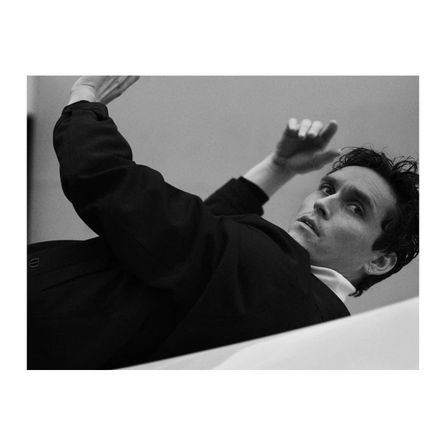 Black and white portrait of a male model with dark hair, wearing a dark jacket and white shirt, lying on a white surface, shot from low angle.