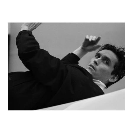 Black and white portrait of a male model with dark hair, wearing a dark jacket and white shirt, lying on a white surface, shot from low angle.
