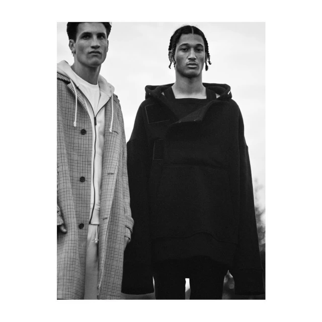 Two adult men stand outdoors wearing hooded sweatshirts and coats. Fashion editorial. Black-and-white photo. Cloudy sky background.