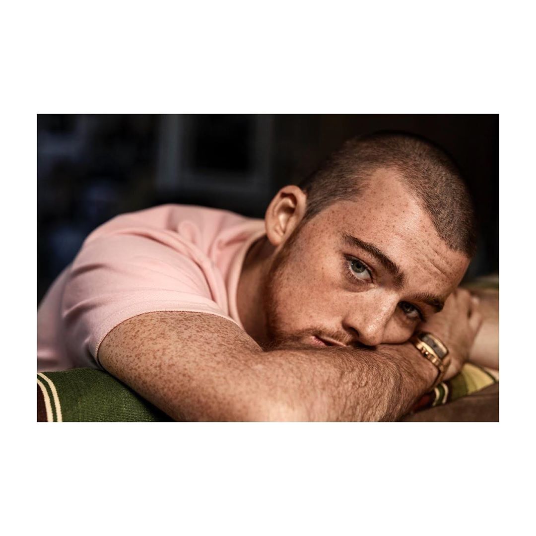 Portrait of an adult male resting his head on his arms, wearing a pink t-shirt, with a shallow depth of field and indoor setting.
