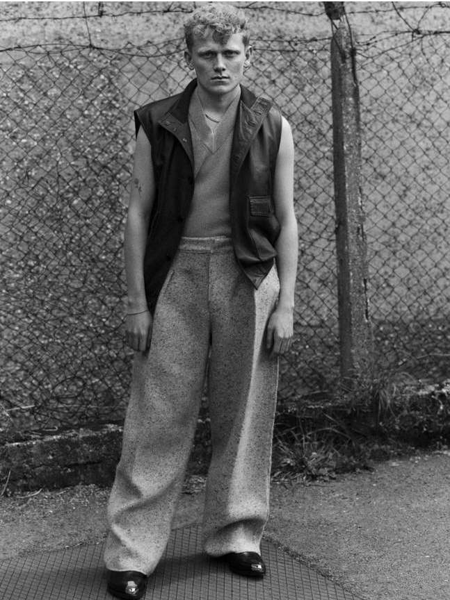 Monochrome portrait of a young man wearing a sleeveless leather vest and textured trousers standing outdoors by a chain-link fence