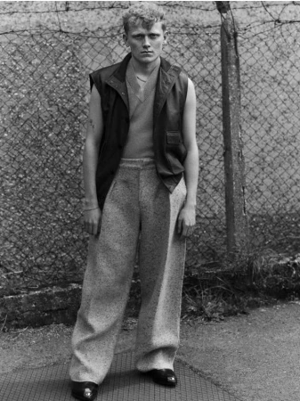 Monochrome portrait of a young man wearing a sleeveless leather vest and textured trousers standing outdoors by a chain-link fence