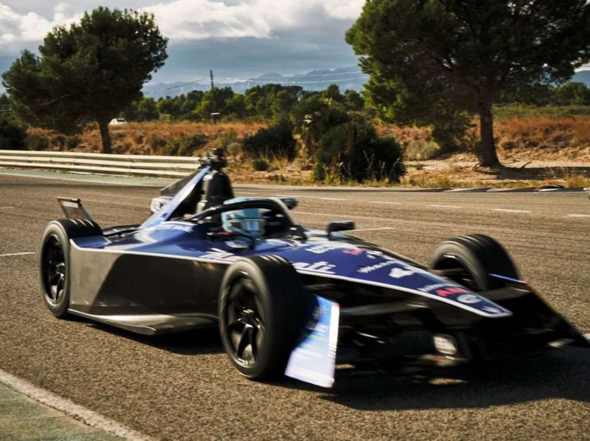 Electric race car speeds along the track during the day, surrounded by lush green trees and vibrant foliage.
