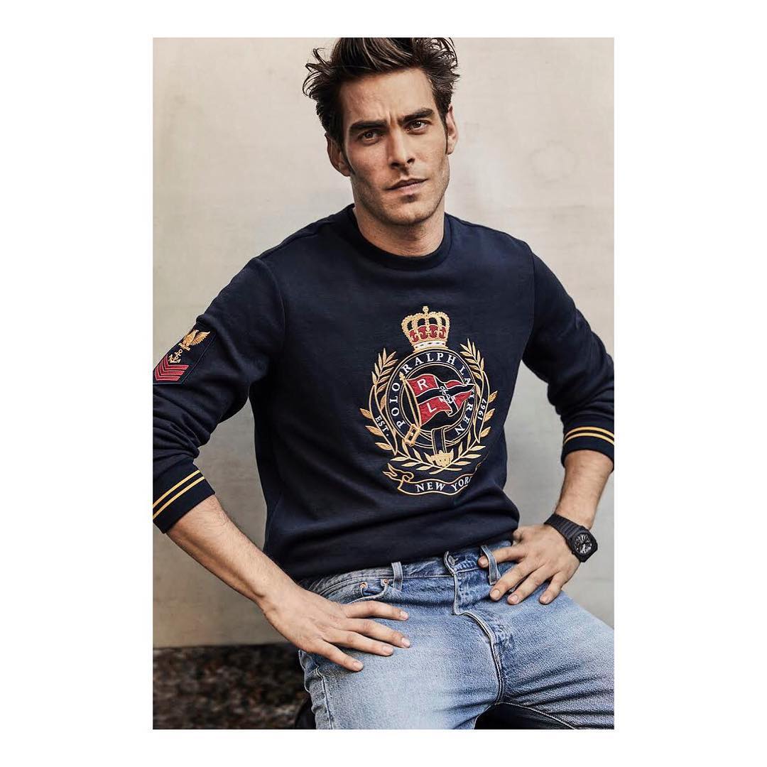A brunette male model poses in front of a grey wall, wearing a navy blue sweater with an ornate crest and light blue denim jeans.