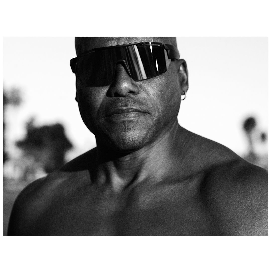 Black and white portrait of a muscular adult male with sunglasses and earring standing outside with trees and sky in the background.