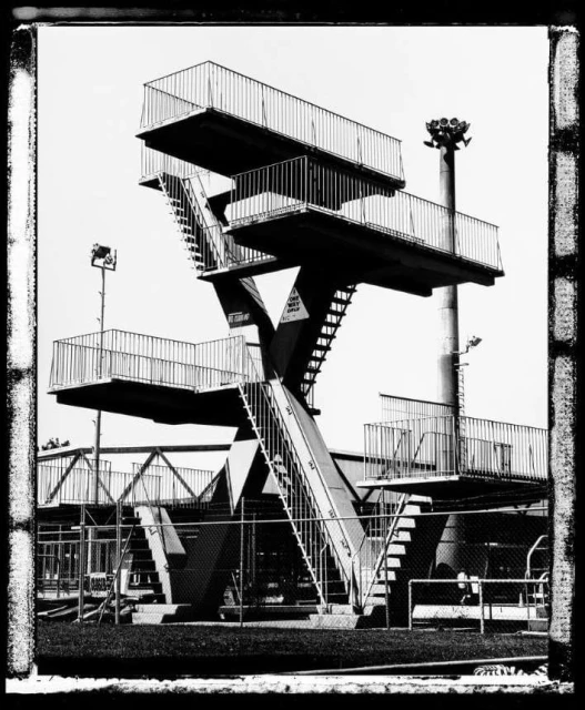 Monochrome photograph of a swimming pool diving platform with a ladder, multiple decks, and metal railings against a light sky.