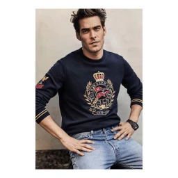 Man wearing a navy sweatshirt with a crest and light wash blue jeans against a neutral wall