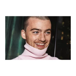 Close-up portrait of Angus Cloud smiling while wearing a pink turtleneck sweater against a dark interior background.