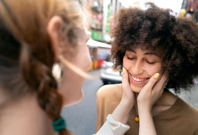 Close-up portrait of two women outdoors, one cupping the other woman's smiling face in a tender, intimate gesture.
