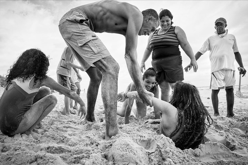 Black and white shot of group on beach digging girl into the sand with overcast sky in the background on a cloudy day.