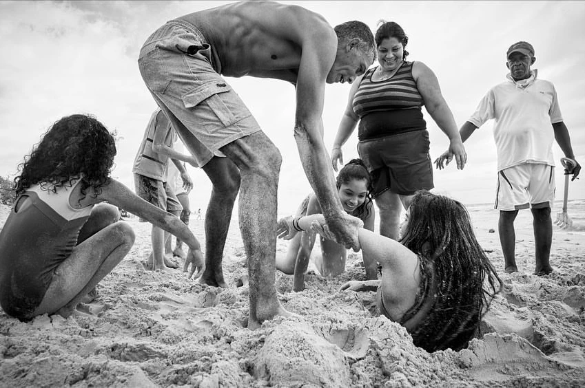 Black and white shot of group on beach digging girl into the sand with overcast sky in the background on a cloudy day.