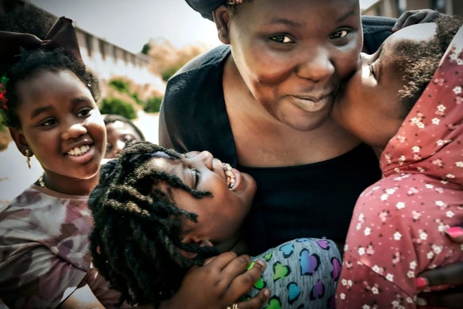 Close-up of a smiling black woman surrounded by four children hugging and kissing her cheek in an outdoor setting.
