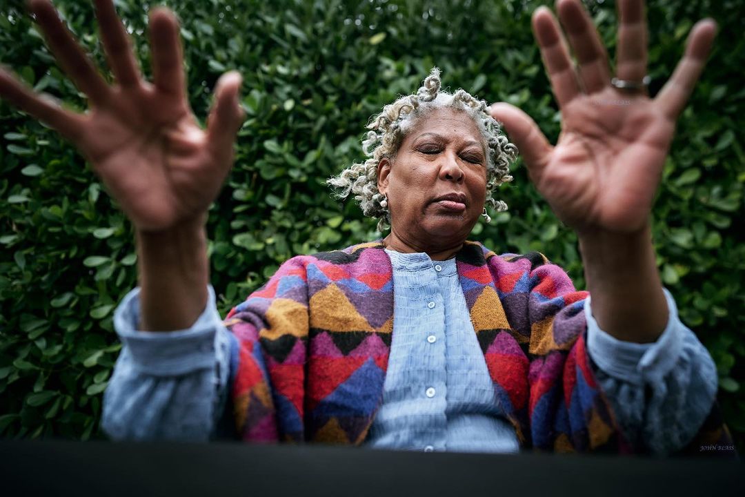 A medium shot of a woman with gray curly hair, wearing a colorful jacket, with her hands raised against a green leafy background.
