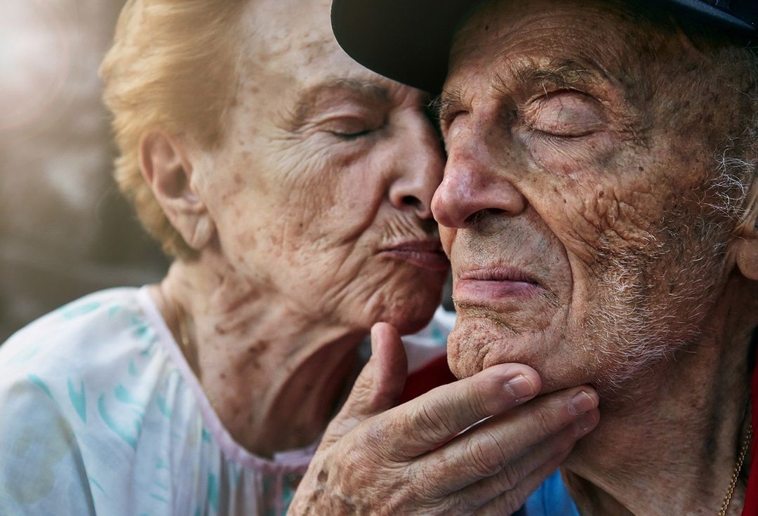 Close-up of an elderly couple with the woman touching the man's face affectionately, both with closed eyes in natural lighting.