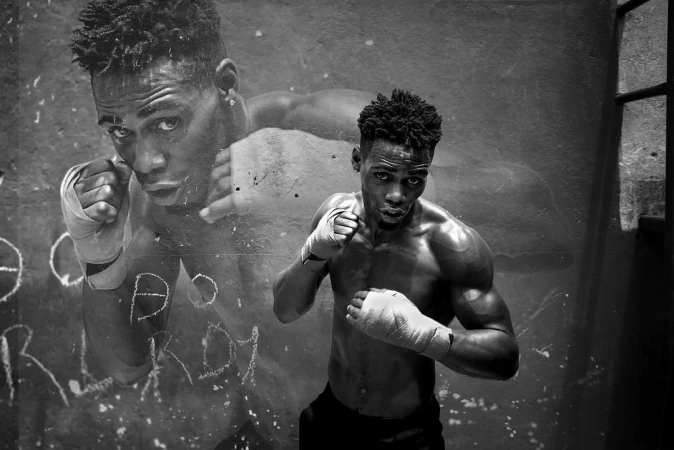 Black and white portrait of muscular African American boxer with hand wraps in a gritty indoor setting, strong pose, intense expression.