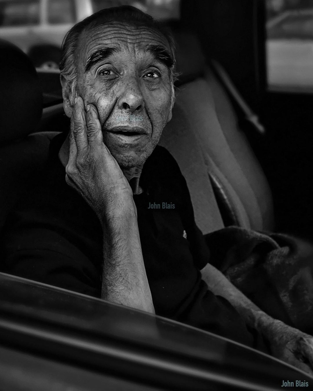 Black and white portrait of an elderly man sitting inside a vehicle with hand on his face in contemplation, with subdued natural lighting.
