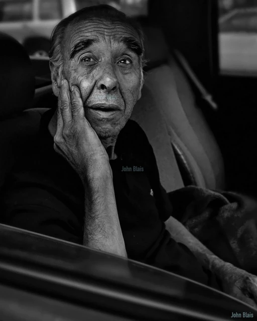 Black and white portrait of an elderly man sitting inside a vehicle with hand on his face in contemplation, with subdued natural lighting.