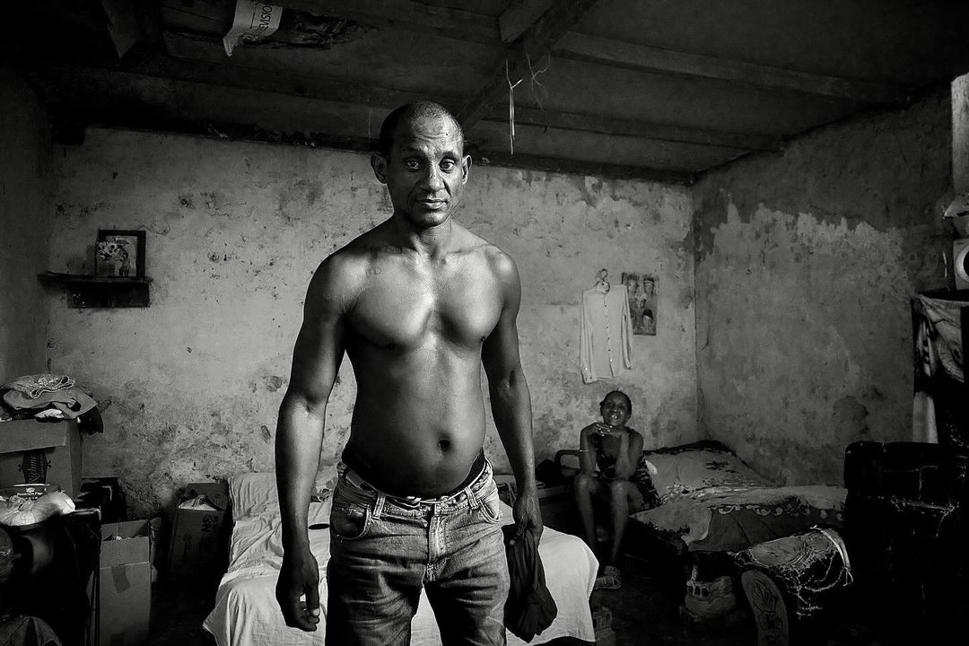 Striking black and white portrait of a shirtless adult male in jeans, standing in an interior setting, with a child sitting in the background.