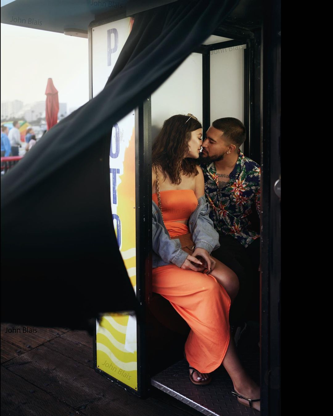 A couple in a photo booth, sharing a kiss, with the woman in an orange dress and the man in a floral shirt in an outdoor setting.