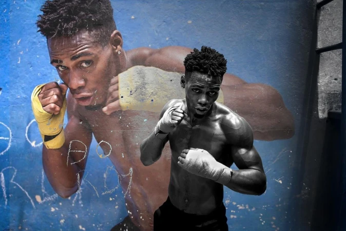 Monochrome portrait of a muscular boxer in fighting stance with hand wraps, over a distressed chalk drawing of another boxer on a blue wall.