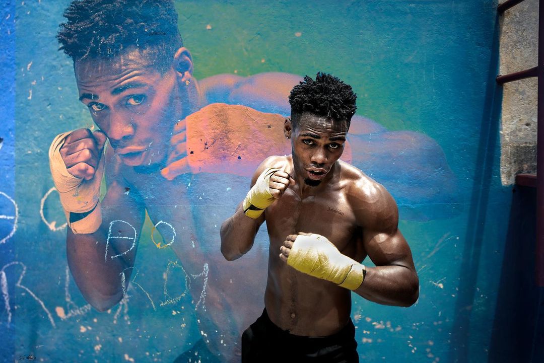 A muscular African-American male boxer stands against a blue graffiti wall with yellow hand wraps, with a mural of the same boxer behind him.