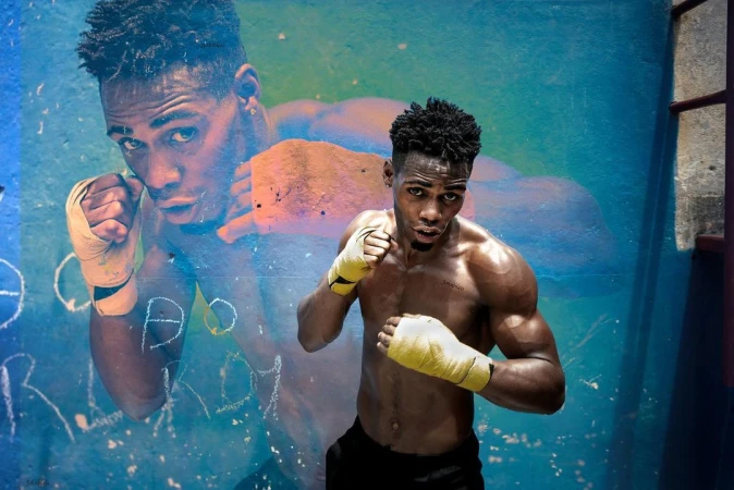 A muscular African-American male boxer stands against a blue graffiti wall with yellow hand wraps, with a mural of the same boxer behind him.