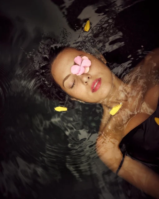 Woman floating in dark water adorned with flower petals. Relaxing self-care concept featuring a person in dark water with flower decoration.