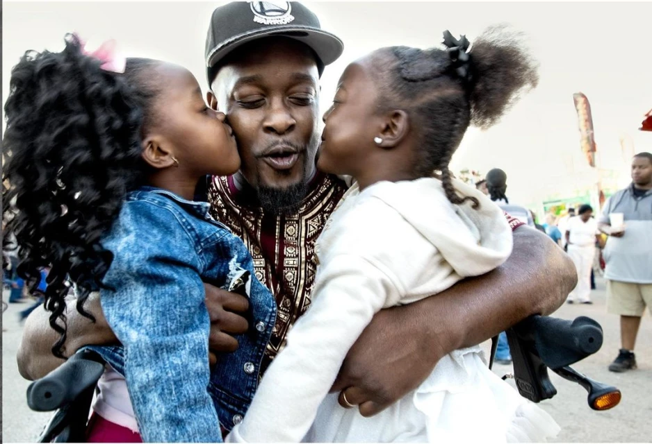 Father in a patterned shirt and baseball cap, embraced by his two daughters, who are kissing him on the cheeks, outdoors.
