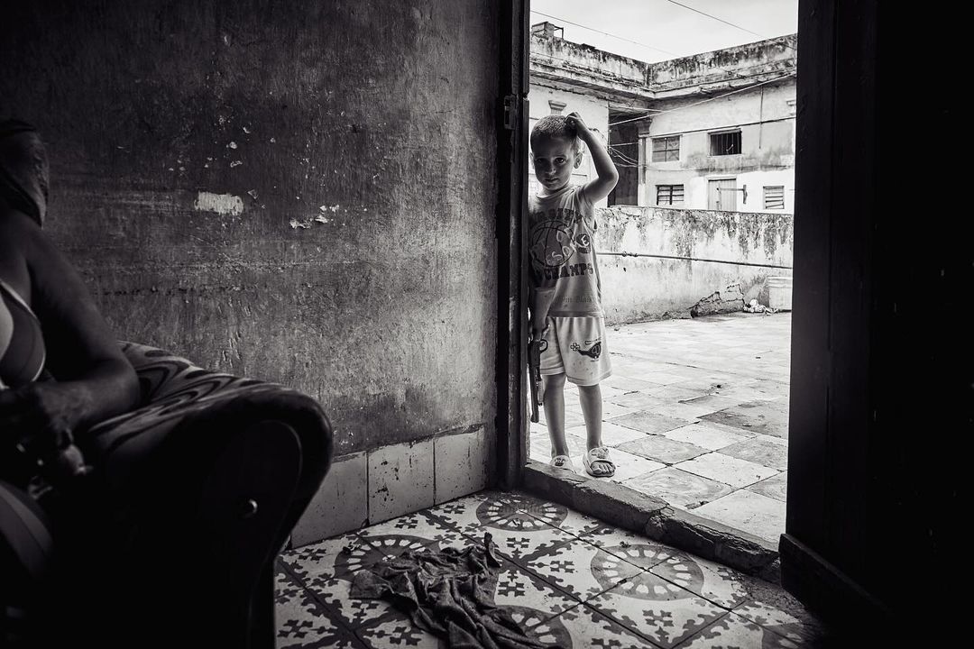 Black and white photo of a child standing in a doorway, looking out onto a rooftop, with an adult seated in the background indoors.