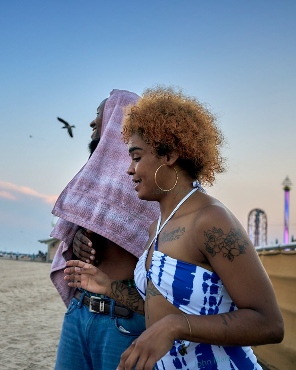 A couple walks on a sandy beach at dusk, the woman with curly hair wears a blue and white top, and the man has a towel over his head.