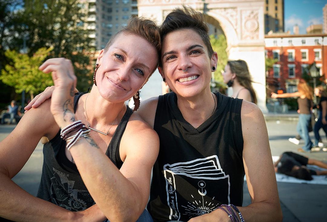 Two women smiling in a sunny urban park, wearing casual attire and showcasing tattoos, with people and an archway in the background.