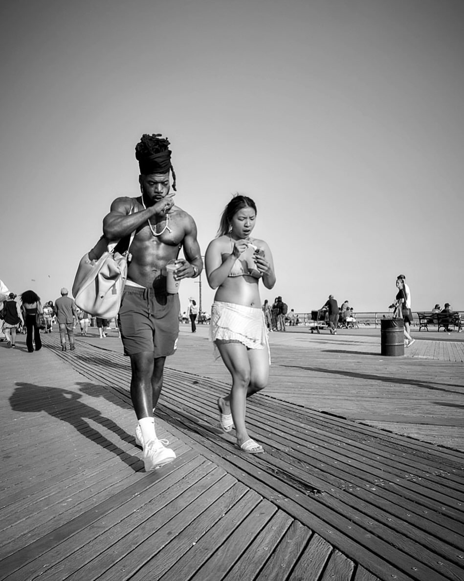 A black and white photo of a muscular man and a woman walking on a boardwalk, both holding beverages, with other people in the background.