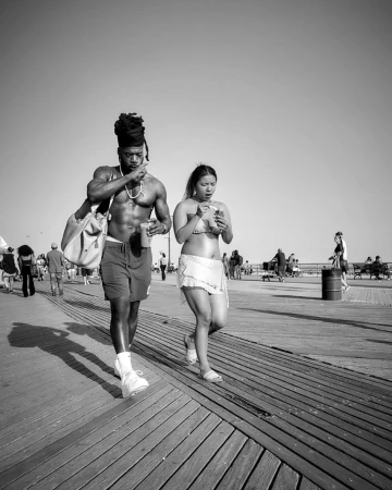 A black and white photo of a muscular man and a woman walking on a boardwalk, both holding beverages, with other people in the background.