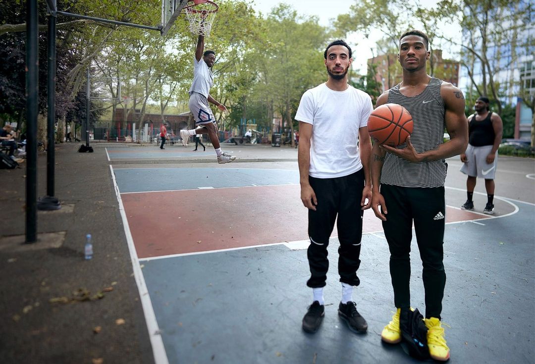 Three adult men on an outdoor basketball court with one athlete doing a slam dunk in daylight against trees and city buildings.