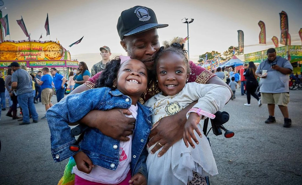 A joyful scene of an African-American man embracing two young girls, all smiling, at a lively outdoor fair with food stands and crowds.