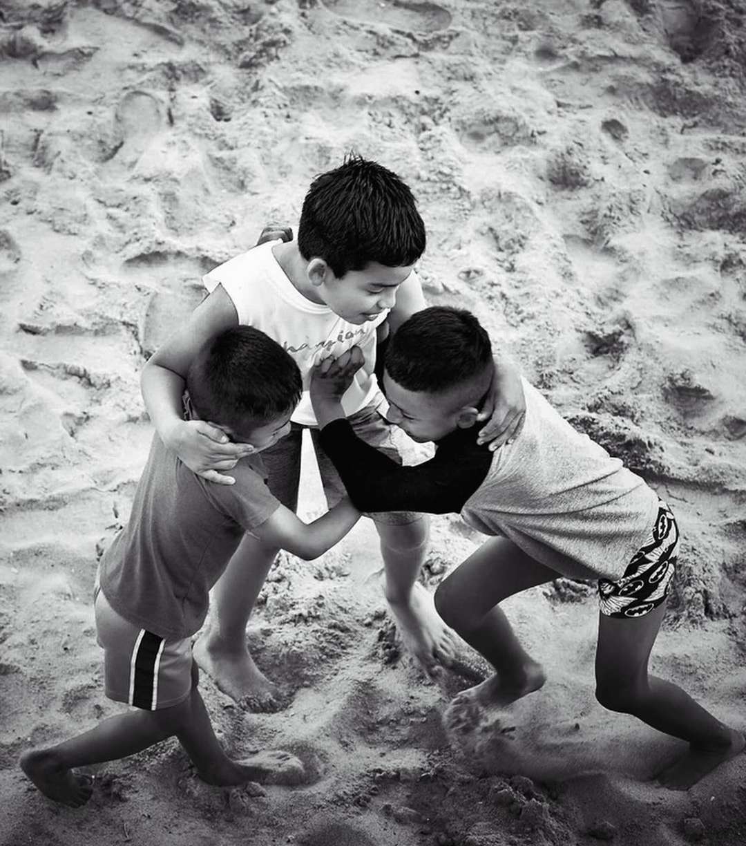 Black and white shot of three young boys playing together on a sandy beach, showcasing childhood friendship and joyful moments.