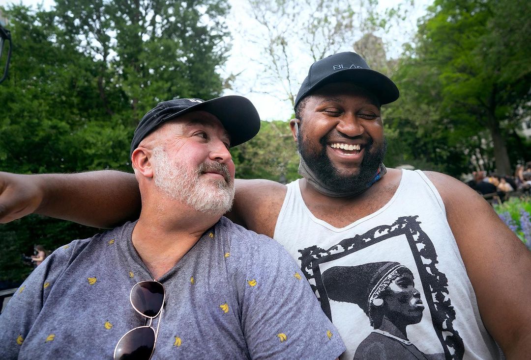 Two men smiling in an outdoor park setting with lush greenery and blooming flowers. They are dressed casually and appear to be in a jovial mood.