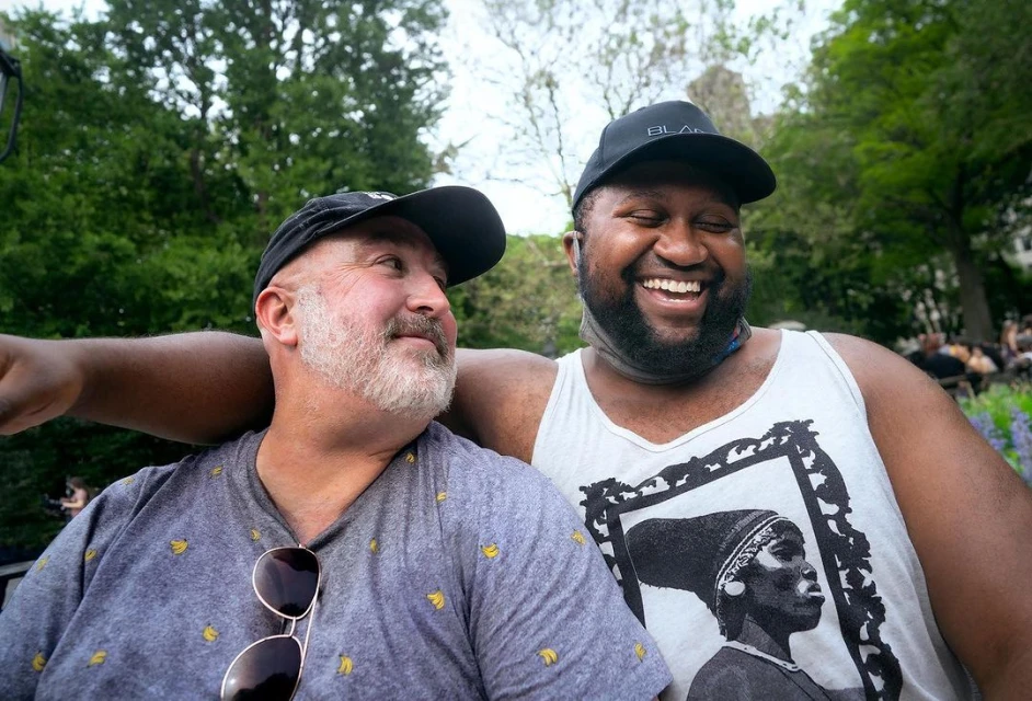 Two men smiling in an outdoor park setting with lush greenery and blooming flowers. They are dressed casually and appear to be in a jovial mood.