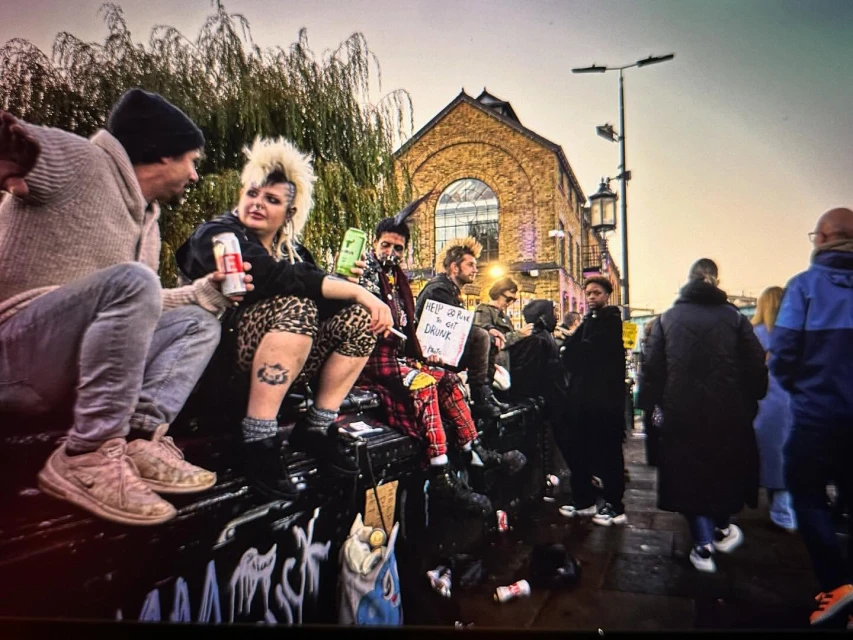 Several people with punk hairstyles sitting outside a brick building with cans and a sign in London, England.