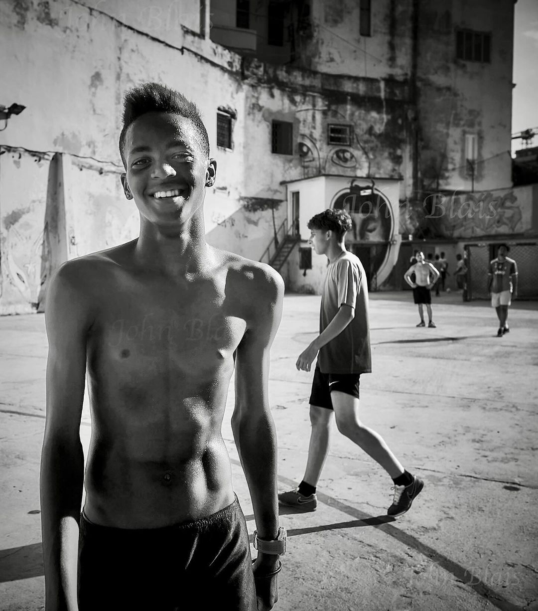 Black and white portrait of a young man smiling in an outdoor court with other people, against a distressed building background.