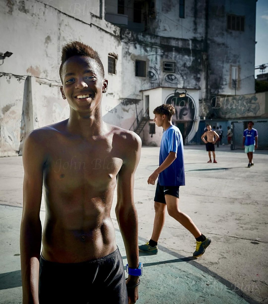 Athletic teenager smiles in outdoor court setting with partial view of basketball game. Old building visible in the background.