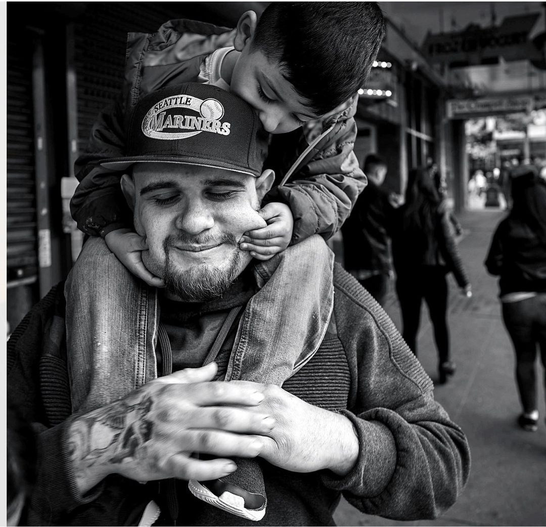 Black and white portrait of a man wearing a baseball cap carrying a small child on his shoulders on a city street.