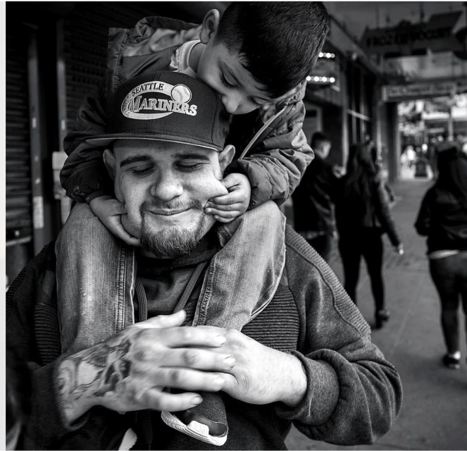 Black and white portrait of a man wearing a baseball cap carrying a small child on his shoulders on a city street.