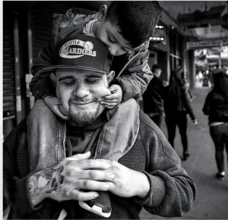 Black and white portrait of a man wearing a baseball cap carrying a small child on his shoulders on a city street.