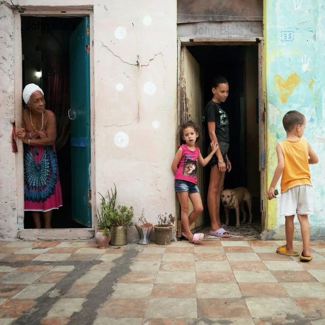 Three children stand near doorways, an elderly woman looks out from another door, a dog is visible in background, checkered floor.