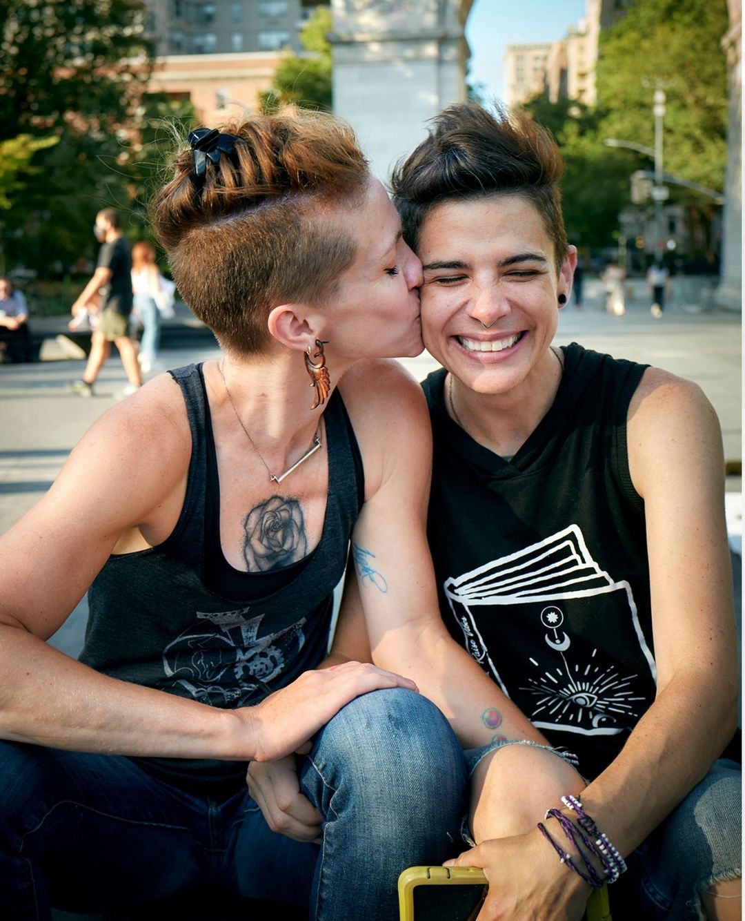 Two women embrace outdoors in a park-like setting with leafy trees. One woman is kissing the cheek of the other, who is smiling.