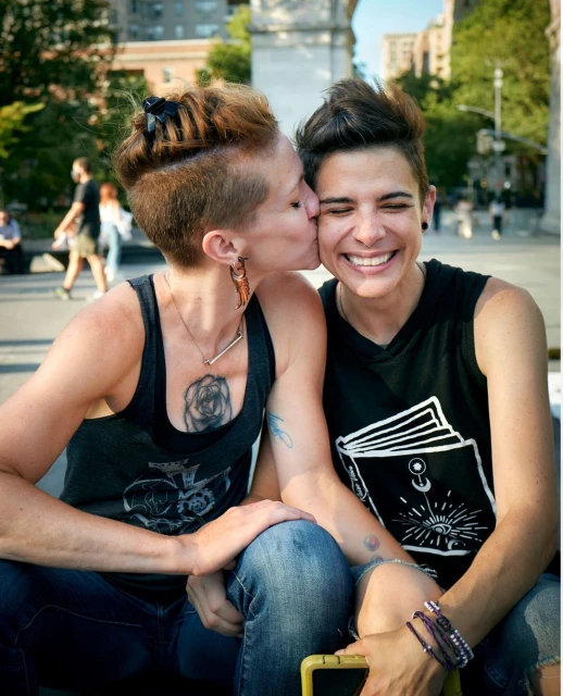 Two women embrace outdoors in a park-like setting with leafy trees. One woman is kissing the cheek of the other, who is smiling.