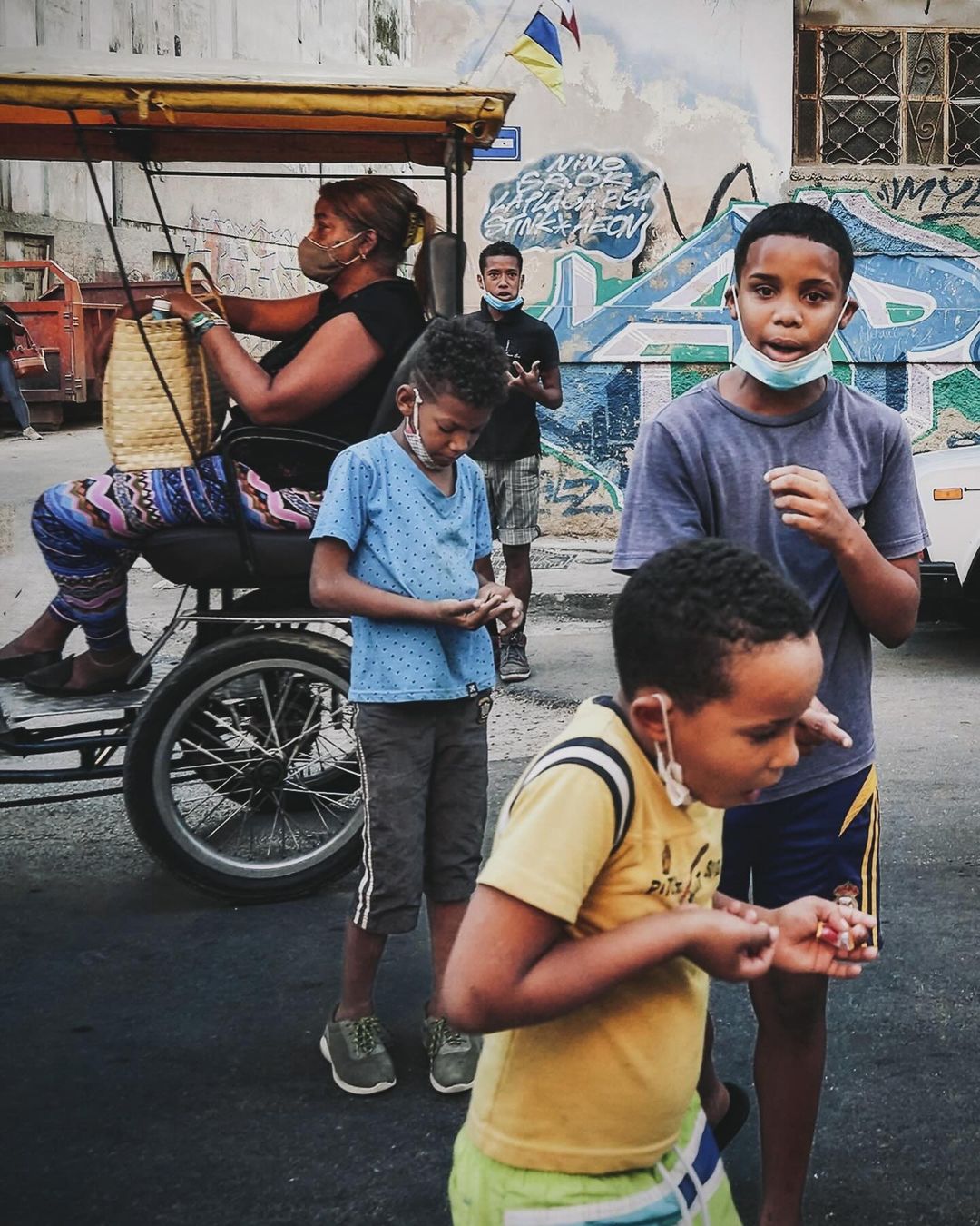 A group of children wearing face masks stand on a city street in front of a woman riding a yellow pedicab with graffiti in the background.